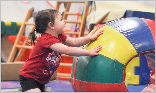 A young participant smiles as she pushes a large, round, colourful ball during a recreational gymnastics class at AIM Gymnastics' Pickering location.